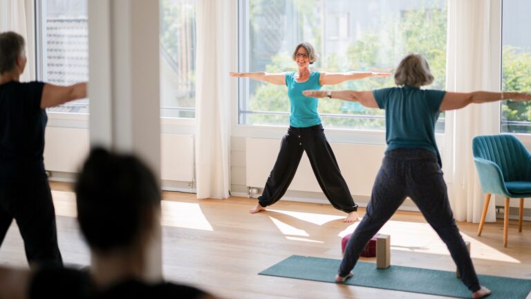 Eine Gruppe von Frauen übt in einem hellen Raum Yoga und folgt den Anweisungen von Yogalehrerin Claudia Benker.