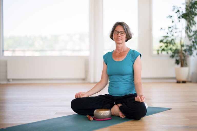 Yogalehrerin Claudia Benker sitzt mit geschlossenen Augen im Schneidersitz auf einer Yogamatte. Vor ihr liegt eine Klangschale.
